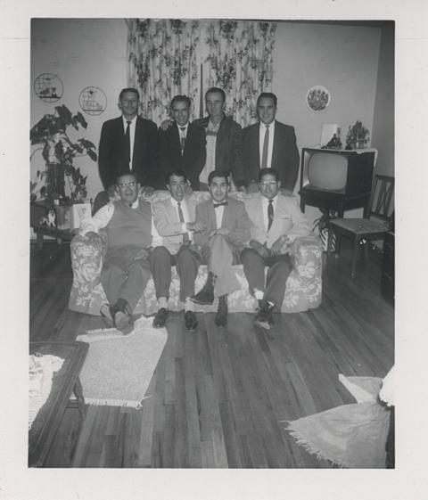 Eight men in coats and ties, four sitting on a sofa and four standing behind them, pose in a residence