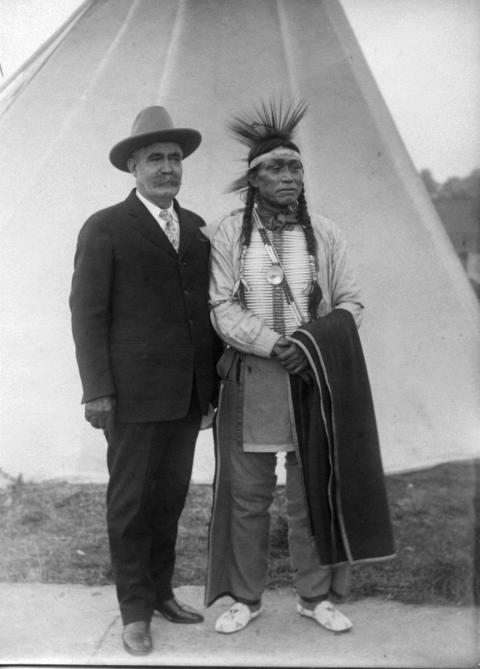 Caucasian man with big moustache, in cowboy hat, suit and tie, stands next to Arapaho man in traditional headband, chest plate and breeches, with a blanket over one arm