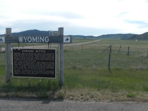 historical marker headlined "Rawhide Buttes" on a roadside with a road leading off into the distance through a treeless plain toward low hills in the distance