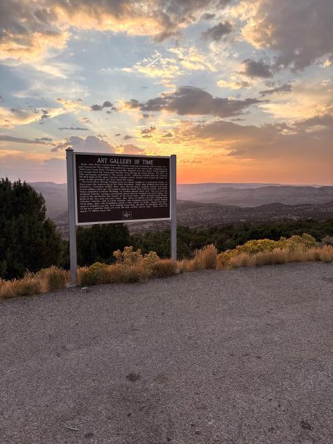 roadside sign title "Art Gallery of Time" perched on the edge of a landscape of rolling hills with exposed strata. The sun is setting in the background.