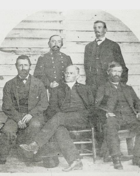 Five men gaze in different directions while posing against a wood slat wall; three in three-piece suits sitting in chairs, with a man in a cavalry uniform ad another in a suit stand behind them 