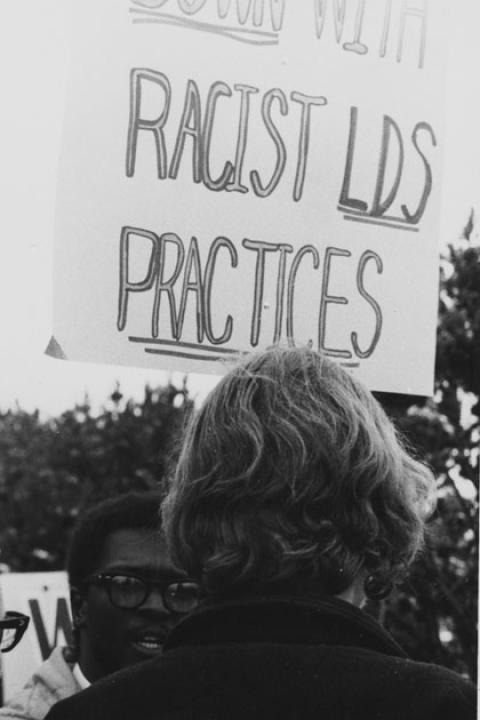A Black man faces another person, seen from behind, holding a sign readiing, "Racist LDS practices"