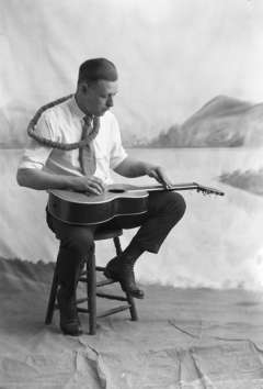 Wyoming musician J. G. Baird plays the steel guitar Hawaiian-style while wearing a lei, 1930s. He plays a standard guitar on his lap with a raised nut to bring strings above the frets. Wyoming State Archives. 